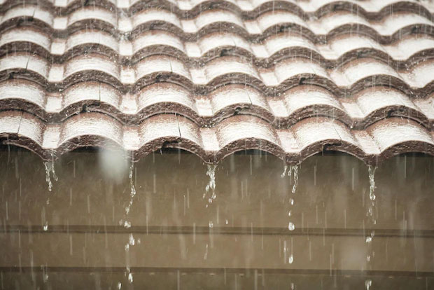IMage shows rain water falling from the edge of a terra cotta tile roof.